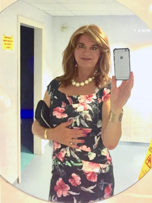 Portrait en miroir d'une personne en robe fleurie tenant un téléphone, avec collier de perles et bijoux, dans un cadre intérieur éclairé.