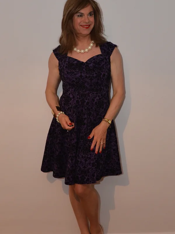 Personne en robe violette à motifs, collier de perles, escarpins beiges, pose debout sur fond clair