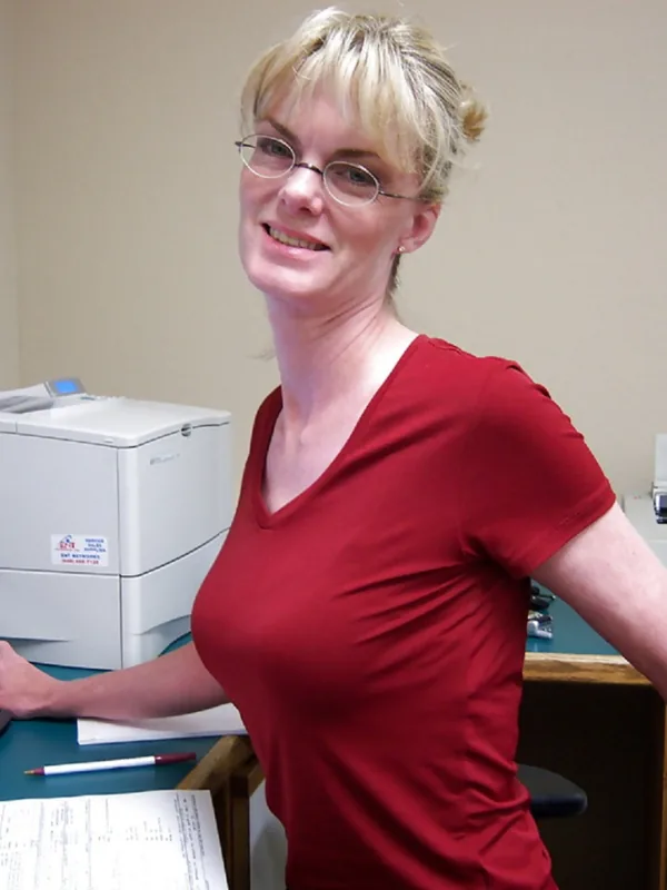 Personne aux cheveux blonds et lunettes, en t-shirt rouge, souriante devant un bureau avec ordinateur et imprimante.