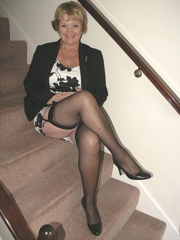 Personne assise sur un escalier, vêtue d'une veste noire, haut blanc à motifs, bas noirs et escarpins, souriant vers l'appareil.