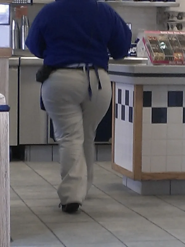 Personne vue de dos en veste bleue et pantalon clair, marchant dans un magasin avec comptoir et étagères.