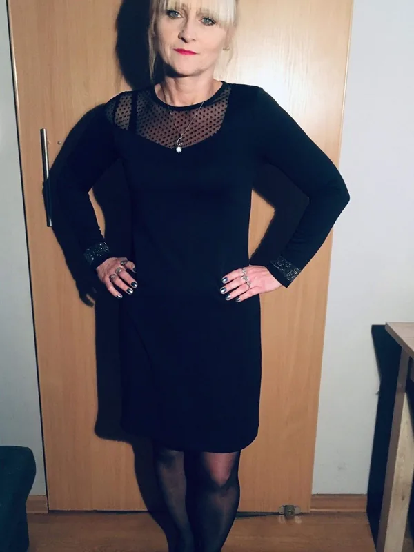 Personne en robe noire à maille, cheveux blonds, mains sur les hanches, devant porte en bois