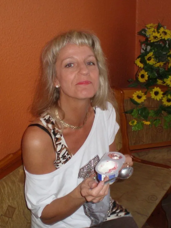 Personne aux cheveux blonds assise, tenant un pot de yaourt, devant un mur orange et des fleurs artificielles