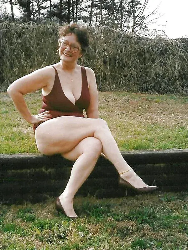 Personne assise sur un banc en bois, portant un maillot de bain rouge foncé et des lunettes, dans un jardin avec une haie en arrière-plan.