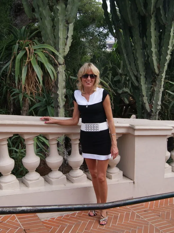 Personne en robe noire et blanche, lunettes de soleil, appuyée sur une balustrade, devant des cactus et plantes vertes.