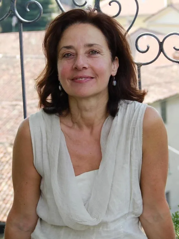 Portrait d'une personne aux cheveux bruns, vêtue d'une robe blanche, devant une grille métallique, sous une lumière naturelle.