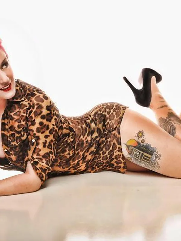 Personne aux cheveux rouges en robe léopard, allongée, tatouages visibles, talons noirs, fond blanc