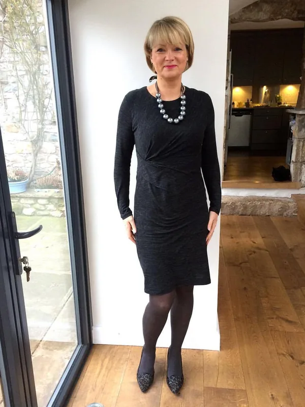 Personne en robe noire, collier de perles, collants foncés, escarpins léopard, debout près d'une porte vitrée dans un intérieur lumineux