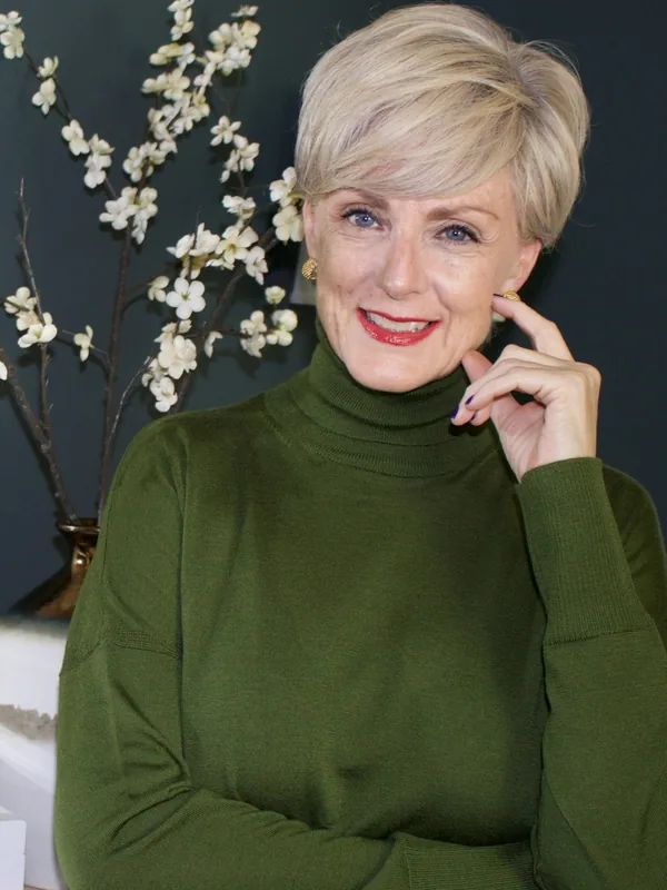Portrait de femme aux cheveux blonds courts, souriante, en pull vert foncé, devant un vase de fleurs blanches sur fond sombre.