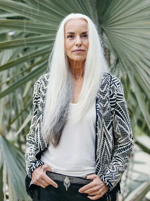 Personne aux cheveux blancs longs, vêtue d'une veste à motifs, devant des feuilles de palmier