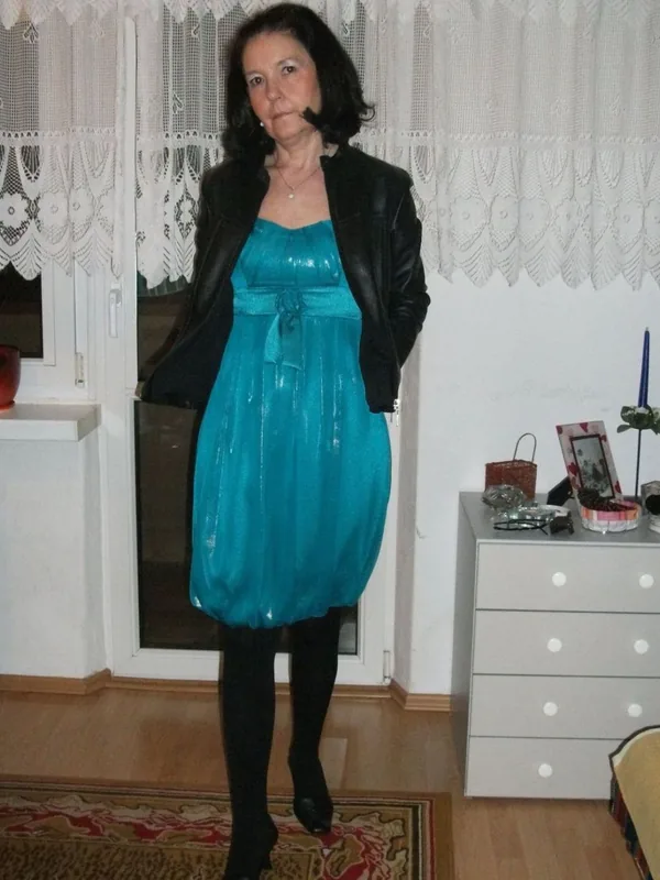 Personne en robe turquoise et veste noire, debout dans une pièce avec rideau dentelle et meuble décoré