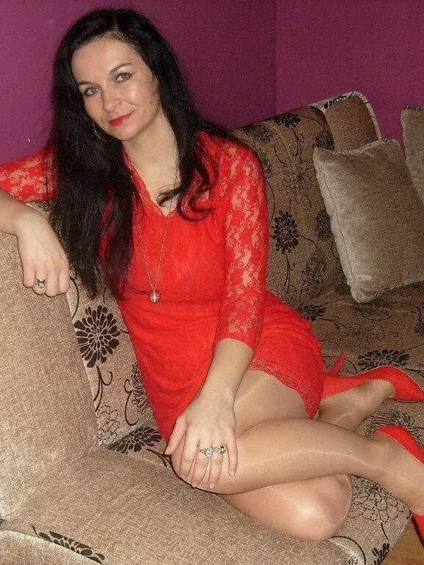 Personne en robe rouge en dentelle, assise sur canapé floral, chaussures rouges, mur violet