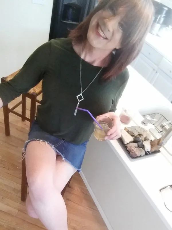 Personne assise en cuisine, tenant un verre avec paille, vêtue d'un haut vert et d'une jupe en jean courte